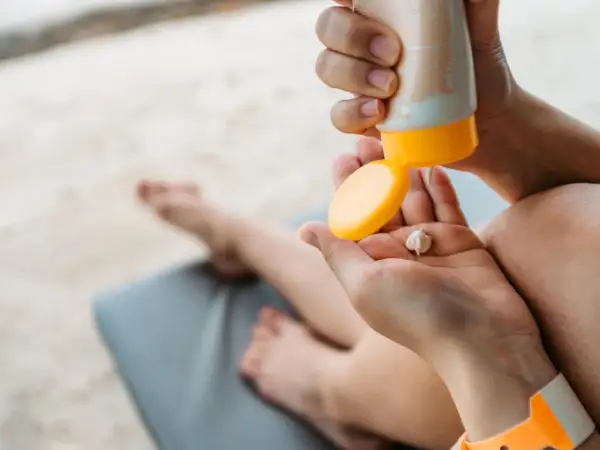 Manos aplicando crema solar caduca en la palma. Un niño descansa en una tumbona al fondo.